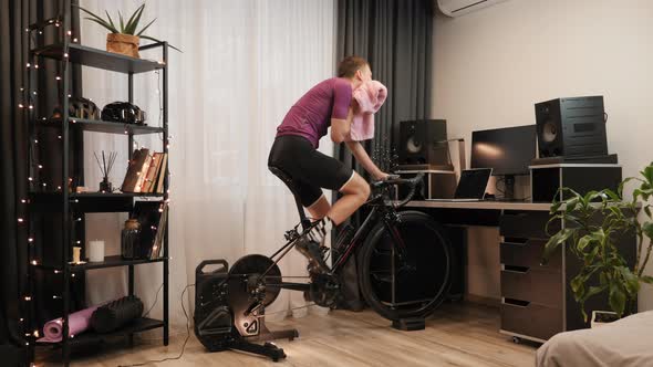 Cyclist is training on exercise bike at home, wiping face with towel during workout