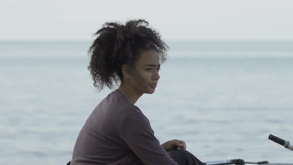 Sad Afro-American Girl Sitting Near Lifeboat on Sea Coast, Shipwreck Victim