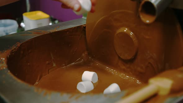 Worker dipping marshmallow with fork in chocolate blending machine