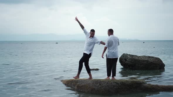 A Joyful Gay Couple Has Fun Dancing on a Rock in Shallow Water Holding Hands with Laughter During