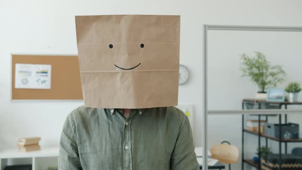 Portrait of Male Employee Wearing Paper Bag with Smile Emoji Standing in Office and Looking at