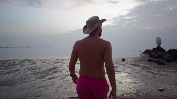 A Shirtless Man in Swim Shorts Walking Towards the Sea at Sunset on Cloudy Day  Back Following Shot