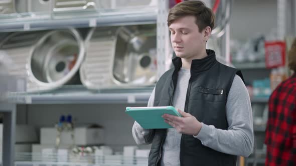 Young Caucasian Worker Checking Goods on Shelves Using Tablet in Hardware Store As Client Passing at