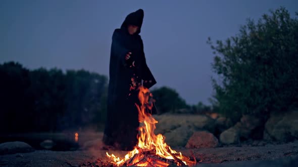 Witch evil in black gown near fire at night. Mysterious wizard doing shamanism over the flame. Scary