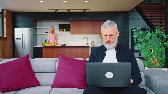 Selective Focus of Serious Adult Male Freelancer Working on Laptop in Kitchen Studio Near His Wife