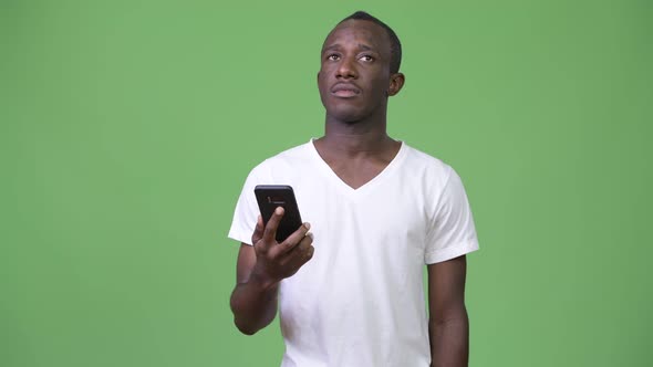 Young African Man Thinking While Using Phone Against Green Background