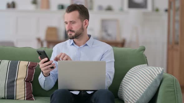 Young Man Working on Smartphone and Laptop on Sofa