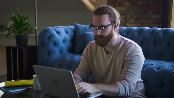 American Bearded Guy Hipster is Working with Laptop While Sitting at Table in Home Interior Spbd