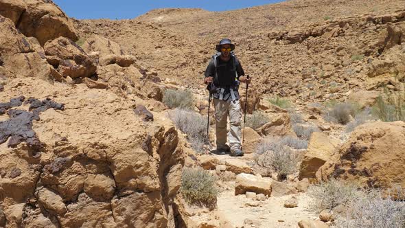 Male Hiker walking in the desert with poles, hat, sunglasses and backpack looking to camera in slow