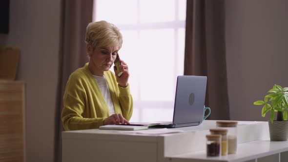 Old Businesswoman is Working in Home Office Communicating By Phone Sitting at Kitchen and Using