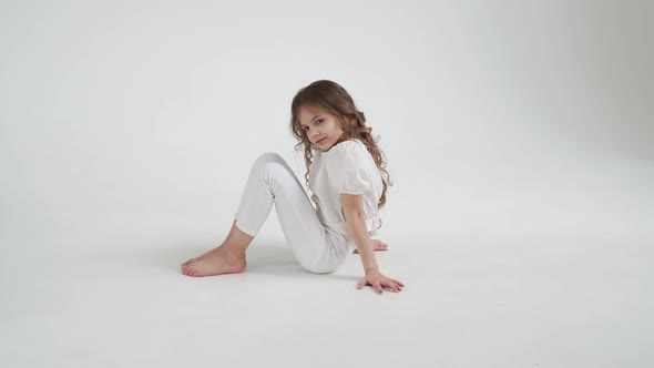 Beautiful Little Girl with Long Hair in White Clothes Sits on the Floor