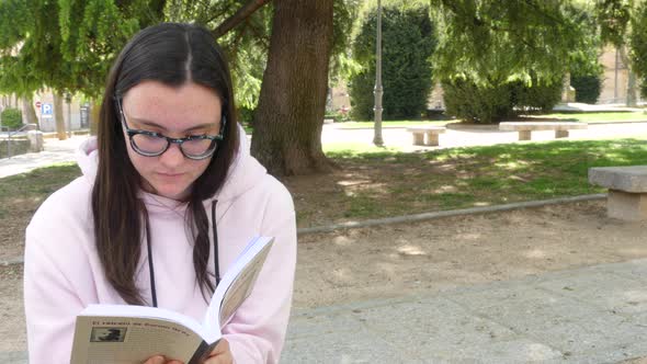 Cute girl reading a novel on the street, next to a park.