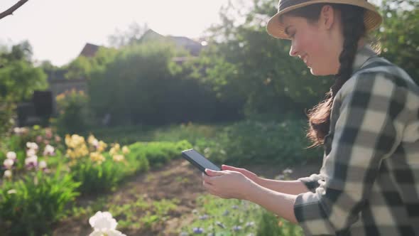 A smiling young woman in a straw hat takes photos of blooming irises on her smartphone