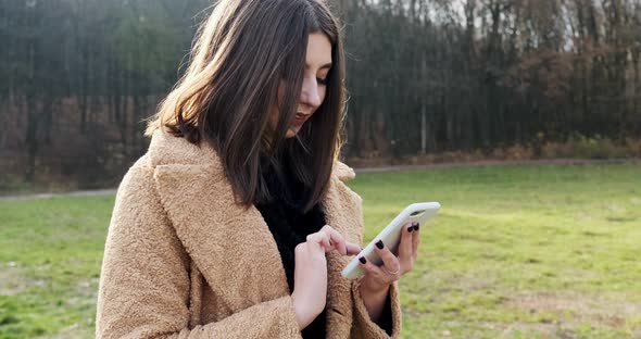 Portrait of Young Attractive Woman Smiling and Looking at Smartphone on the Green Lawn at Autumn