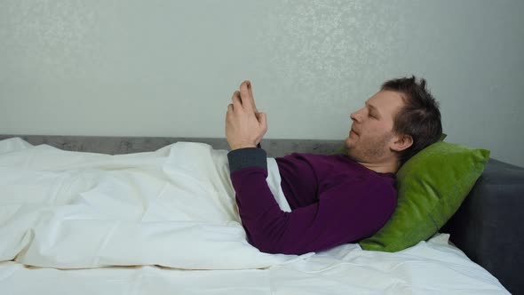 Man Covered with a Blanket is Lying on the Bed and Playing a Mobile Game