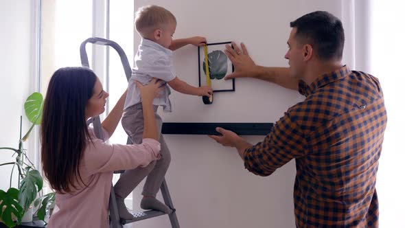 Pretty Kid with Tape Measure Helps Mother and Father Hang Shelf with Picture on Wall After Repair in