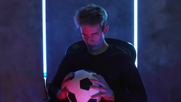 Soccer Player Holds Ball in Hands Sits on Chair and Looks at Camera Against Bright Lights in Dark