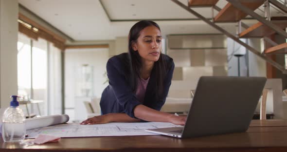 Mixed race woman by desk at home using laptop looking through blueprints