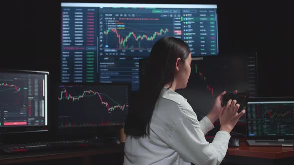 Asian Female  Stock Market Broker Using Mobile Phone With Analysing Graphs On Multiple Computer