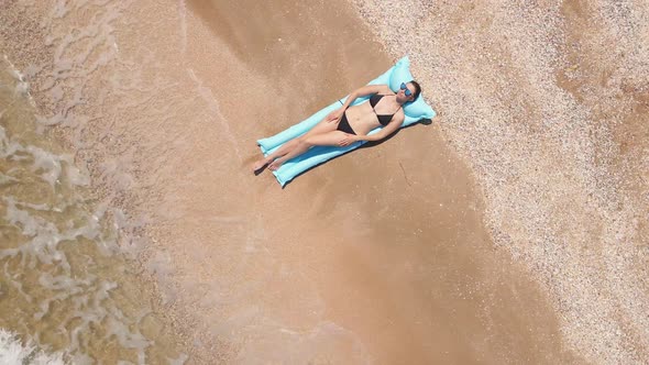 Woman in a Black Swimsuit Lies on a Blue Mattress on a Sandy Beach By the Sea