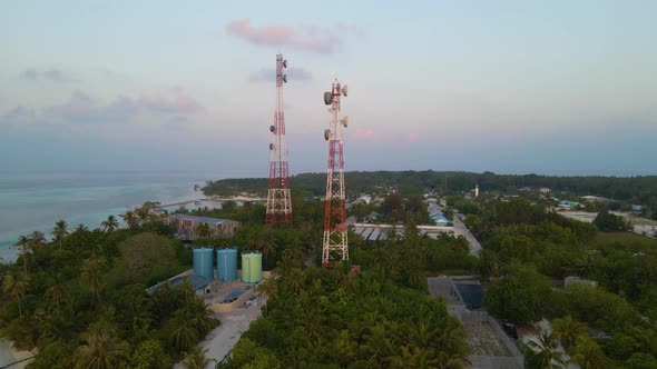 Two cell towers surrounded by green trees and buildings on the shores of the turquoise sea. Maldives