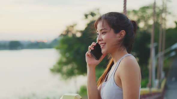 Asian young sport woman using and talking on the telephone with happiness in evening sunset at park