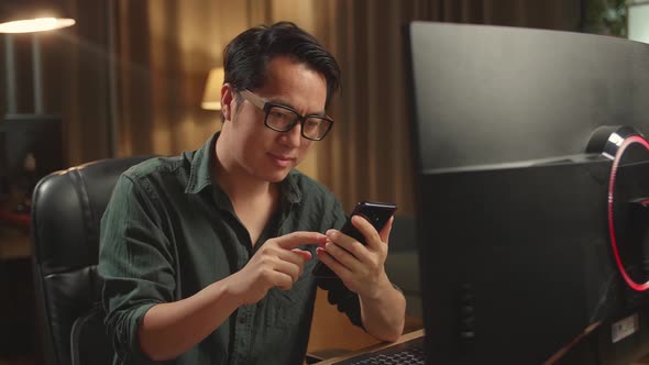Asian Male Using Mobile Phone While Works On His Personal Computer With Big Display