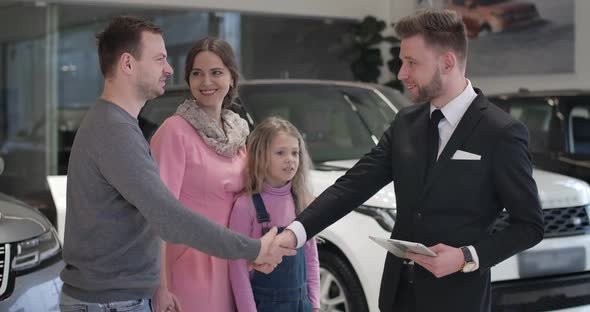 Successful Caucasian Businessman Shaking Car Dealer's Hand and Kissing Wife on Cheek. Cheerful