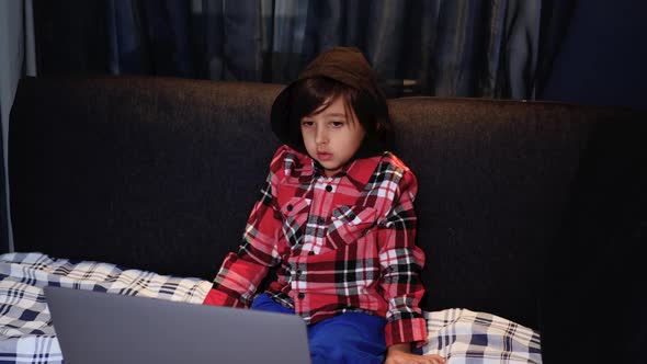 Boy Child in a Red Plaid is Watching a Computer Sitting on Bed at Home