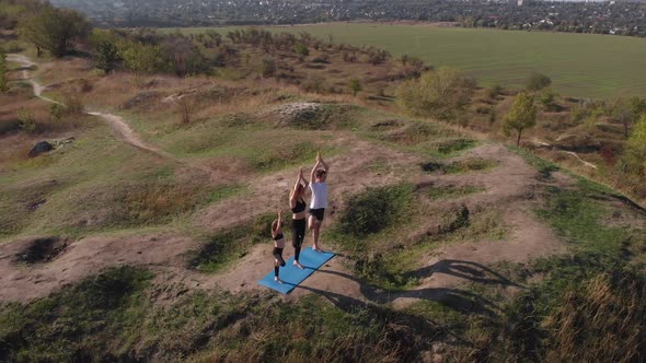 Orbiting Aerial Shot of Family of Three Mother Father and Daughter Do Yoga Exercises on Top of Hill