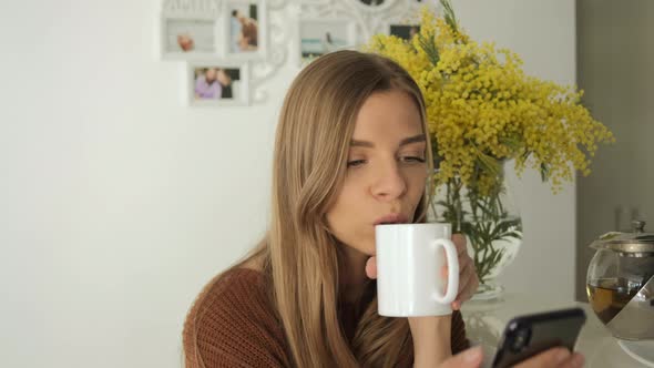 Young blond woman using smartphone and drinks tea in the white kitchen in good mood