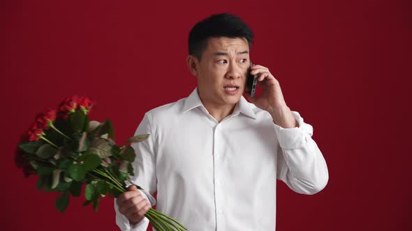 Displeased Asian man holding a bouquet of red roses and talking by phone