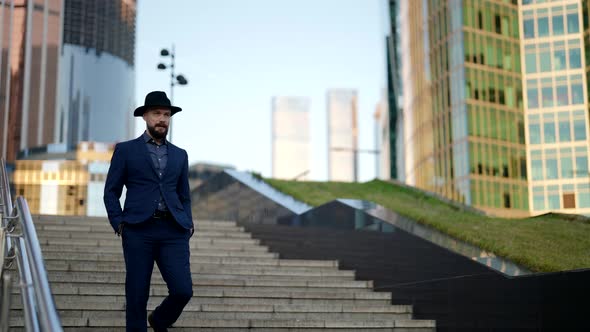 a Bearded and Mustachioed Stylish Man in a Hat and a Plaid Suit Has Hands in Pockets and is Walking