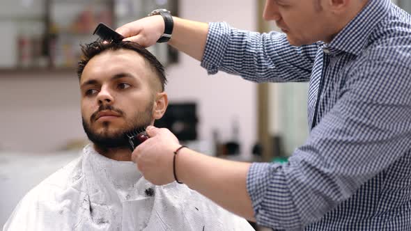 Unparalleled Barber with a Beard and a Tattoo Is Cutting the Hair of His Client in the Barbershop.