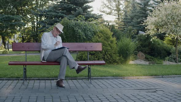 Senior Poet in Hat and Suit Sitting on Park Bench Thinking and Writing