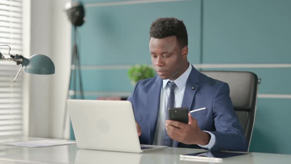 African Businessman Using Smartphone While Using Laptop in Office