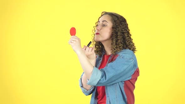 Curly Woman Paints Her Lips Looking in a Red Mirror on Yellow Background, Slow Motion