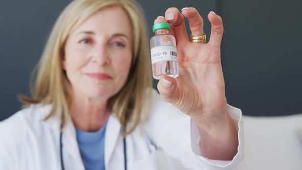 Caucasian senior female doctor holding vial of coronavirus vaccine to camera and smiling