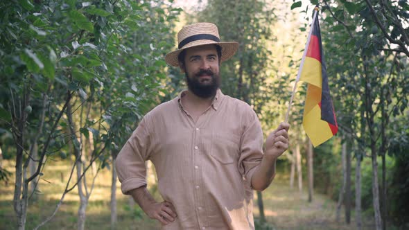 Positive German Bearded Gardener with Mustache in Straw Hat Posing Outdoors with National Flag on