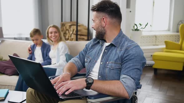 Smiling Disabled Guy with Beard Sitting in Wheelchair and Working on Laptop, Talking With His Wife