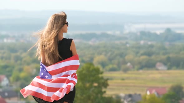 Young Pretty American Woman with Long Hair Holding Waving on Wind USA Flag on Her Sholders Standing