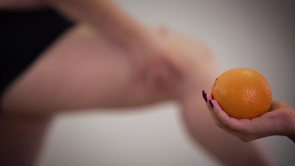 Blurred Caucasian Obese Woman Touching Fat Legs at the Background As Female Hand Holding Orange 