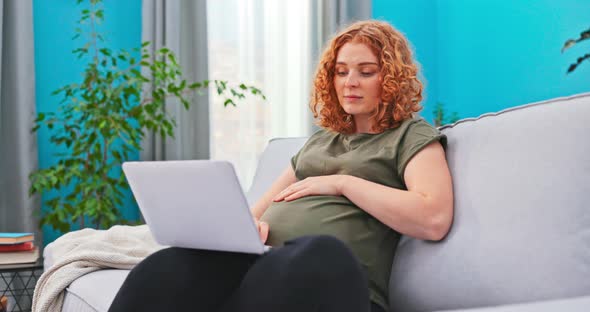 Beautiful Redheaded Young Woman Sits on Living Room Couch with Laptop in Lap Clutching Her