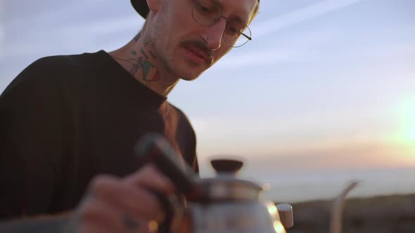 Cool Mustached Guy with Piercing and Tattoo on His Neck Holding Kettle with Boiled Water on Seashore