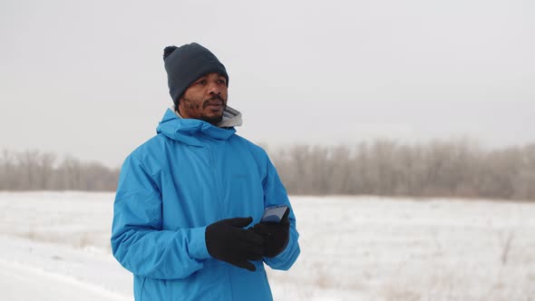 Black Man Stands Outdoor in Winter Clothes and Looks Into Smartphone Using GPS Application Front