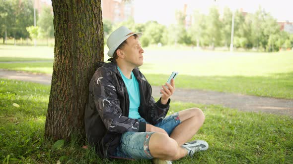 Man with hat on a sunny day sits with a mobile phone under a tree in a city park, camera movement