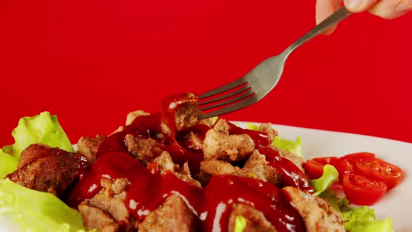 A Man's Hand with a Fork Dips a Piece of Meat in Ketchup on a Red Background