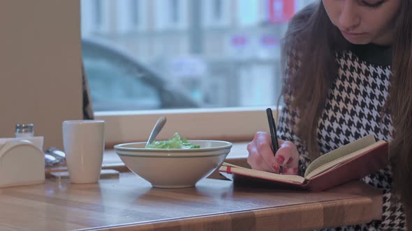 Arms of Young Businesswoman Writing in Note Book. She Is Sitting at the Table in Cafe and Eating a