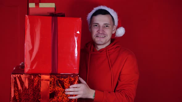 Young Handsome Man in Santa Claus Hat Holds Gifts on Red Background