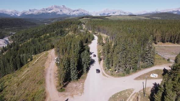 Two cars driving along a dusty logging road through coniferous forests with a majestic Rocky Mountai